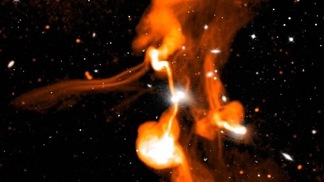 Two giant radio galaxies (more than one million light-years from end to end) at the center of a large group of galaxies in the cluster Abell 194, revealing the presence of relatively narrow magnetic filaments in the region, as well as complex interactions between the radio emission from the two galaxies. The MeerKAT radio image is shown in orange, with an optical image dominated by normal galaxies shown in white. Adapted from K. Knowles et al., “The MeerKAT Galaxy Cluster Legacy Survey. I. Survey Overview and Highlights” (Astronomy & Astrophysics, in press). Image credit: SARAO, SDSS.